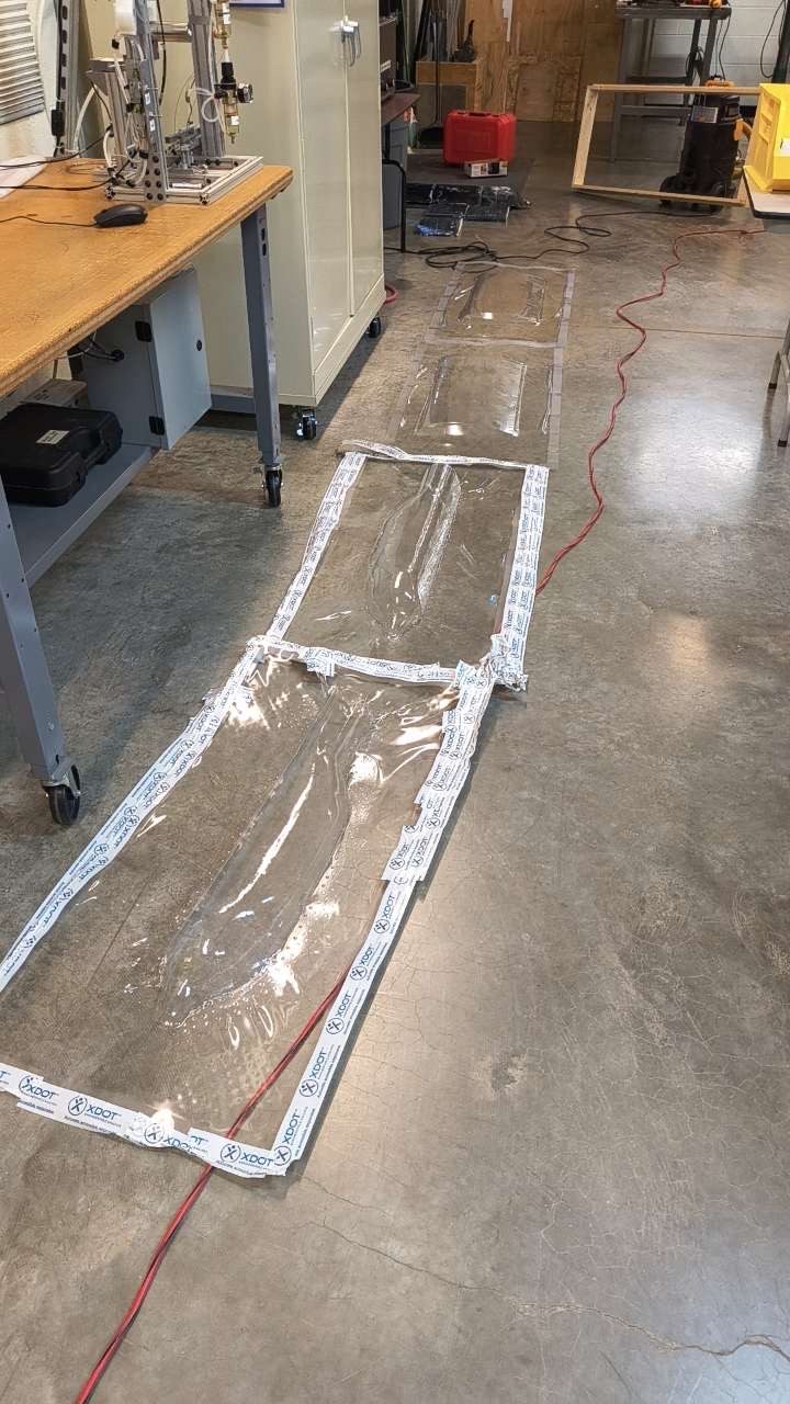 Photo of long clear vacuum-formed plastic skin sheets laid out on a concrete shop floor, edged with white tape.