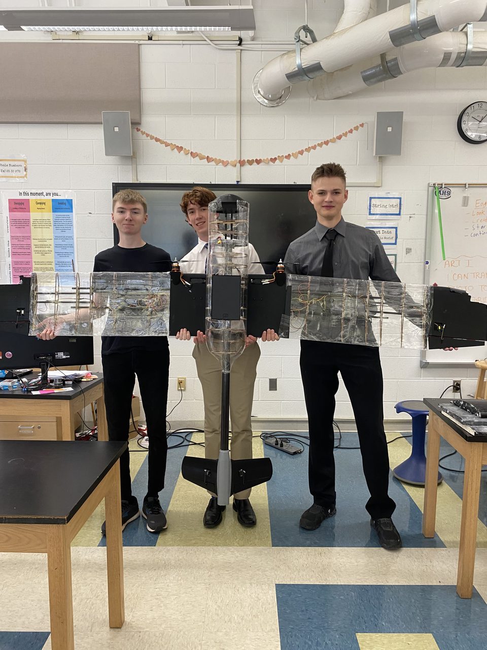 Photo of three students in a classroom holding the completed aircraft horizontally, with its clear ribbed wings and fuselage, black wing center section, twin motors, and black V-tail.