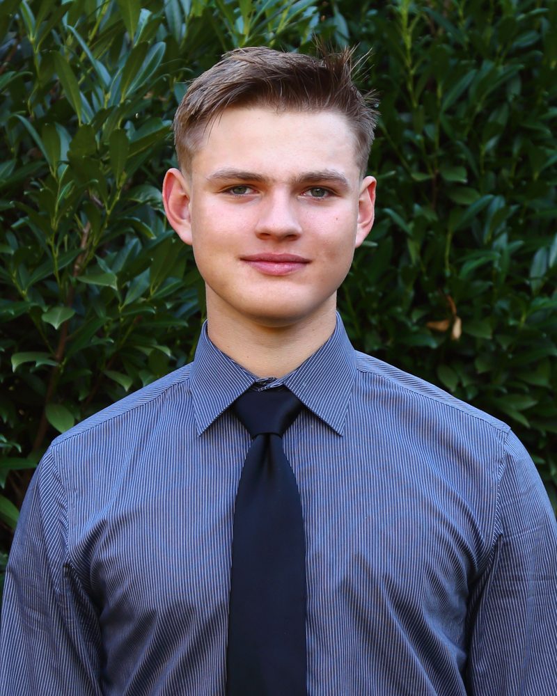 Portrait of Michael Znachonok, facing the camera in a blue-gray pinstriped collared shirt and black necktie, against a dense dark-green foliage background.