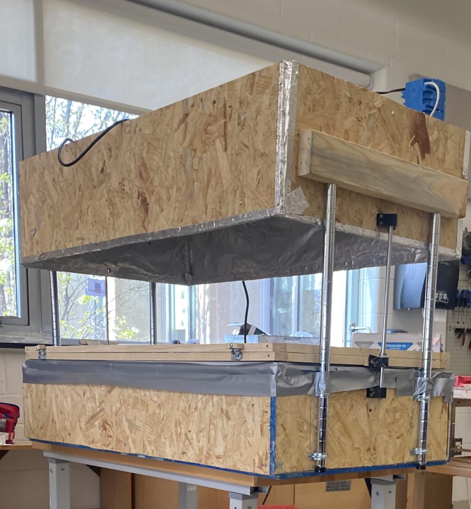 Photo of the full-size vacuum former: a large foil-lined heater box raised on steel posts above a wide framed forming bed with a latched clamping frame.