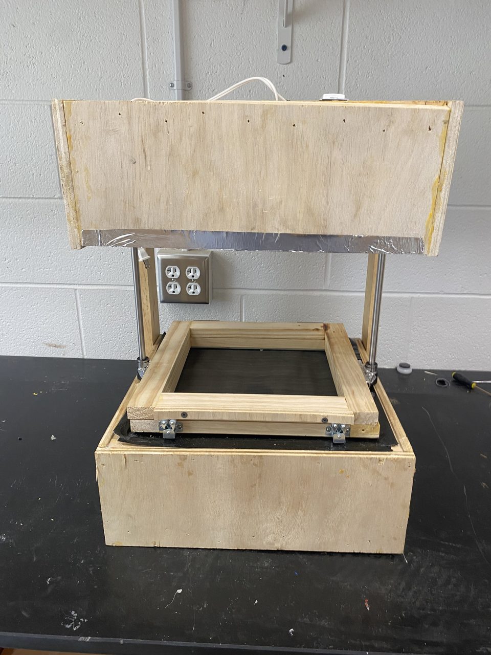 Front photo of the same small vacuum former, showing the foil-lined heater box, the two steel guide rods, the latched wooden clamping frame, and the dark mesh forming surface below.