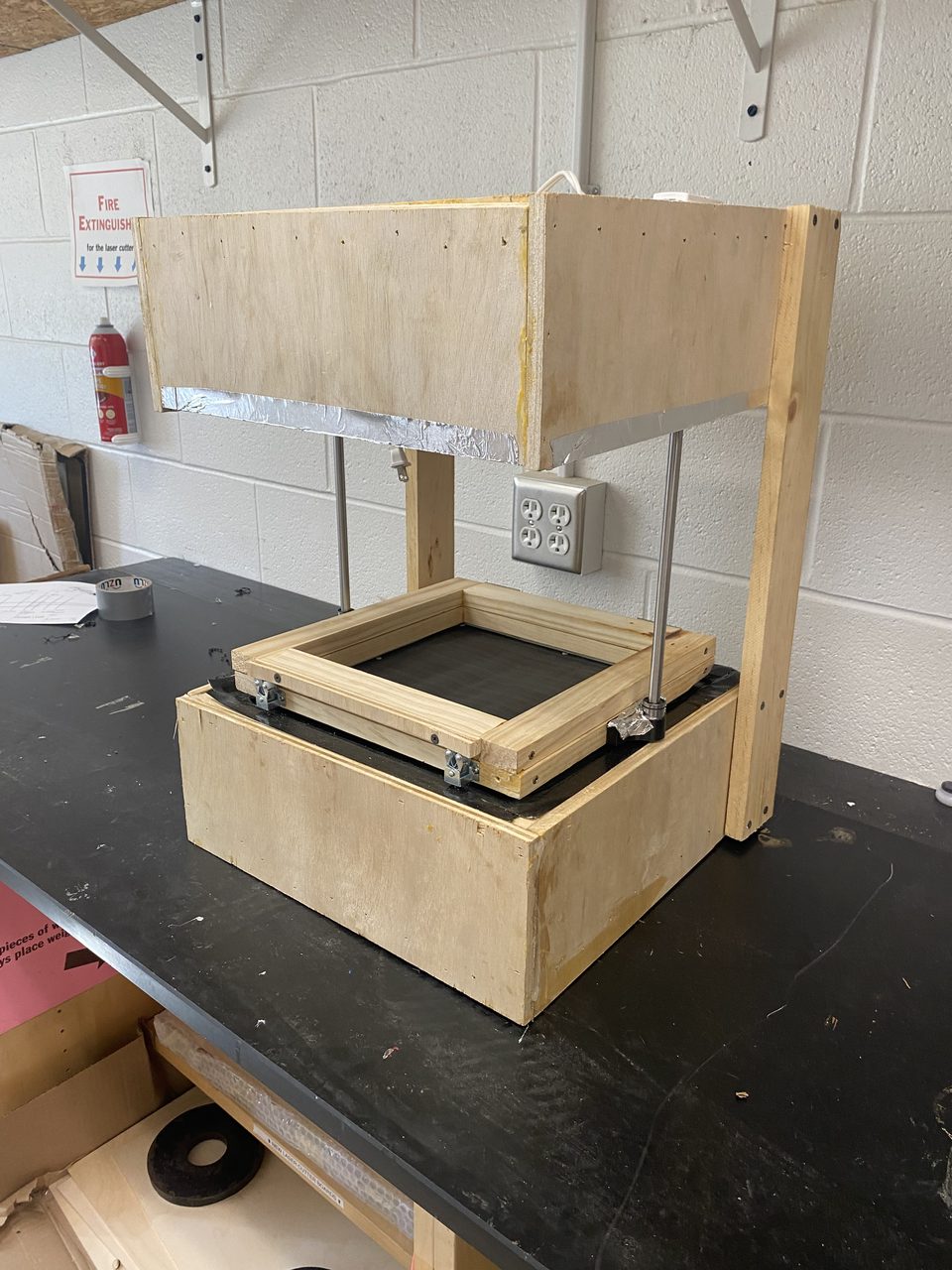 Photo of the small plywood vacuum former on a black lab bench: a heater box lined with aluminum foil held above the base on two steel rods, a square wooden clamping frame with latches, and a dark mesh forming surface on top of the base box.